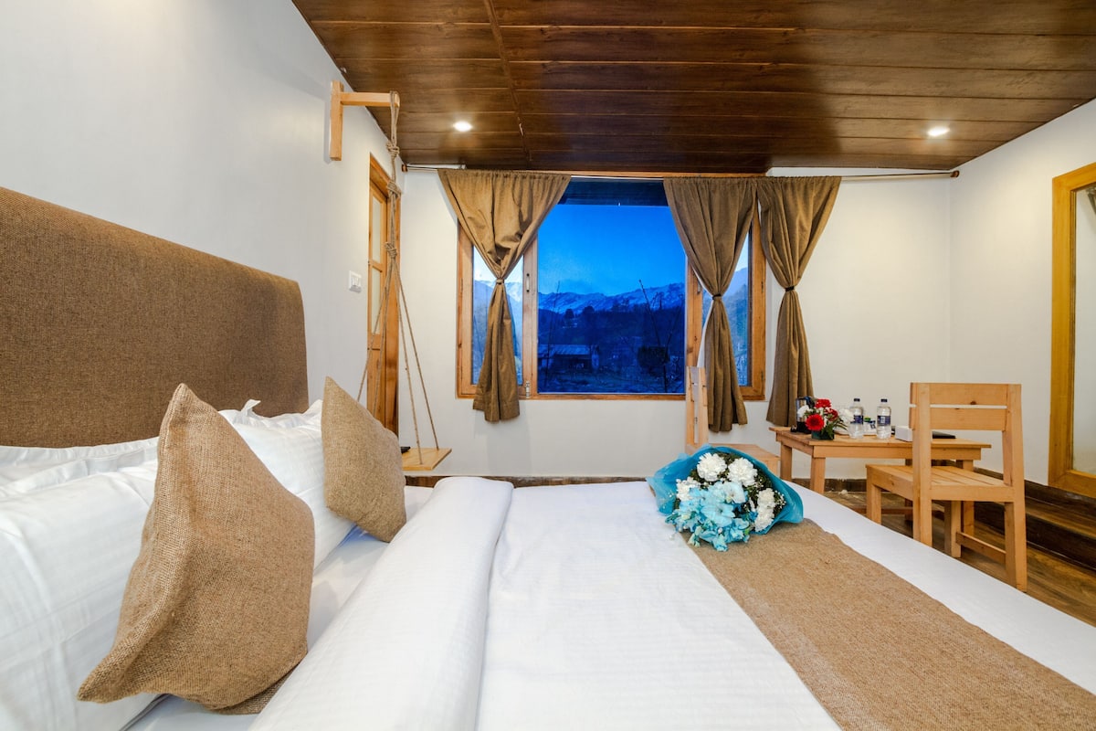 Habitación de hotel con cama, cabecera de madera, cortinas, ventana con vista a la montaña, una silla de madera y una mesa con un ramo de flores.
