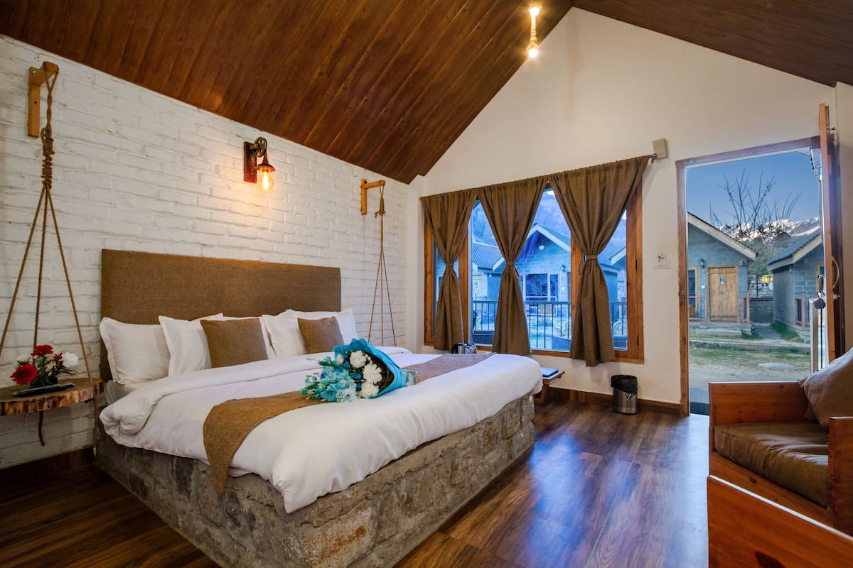 Un dormitorio con cama de piedra, techo de madera, paredes de ladrillo blanco y vista a un edificio exterior.