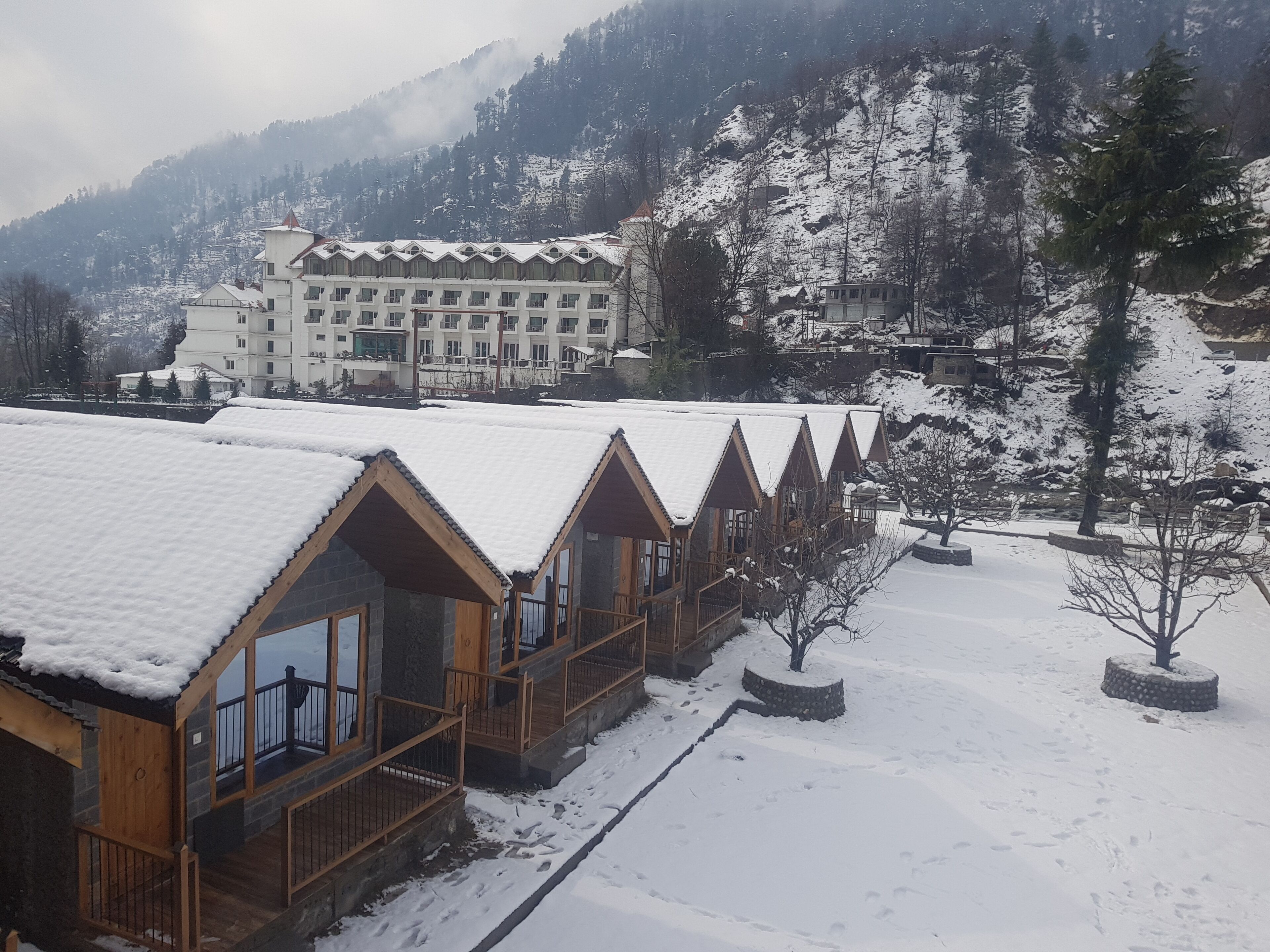 A row of wooden cabins with snow-covered roofs, set against a backdrop of a large hotel and snow-covered mountains.