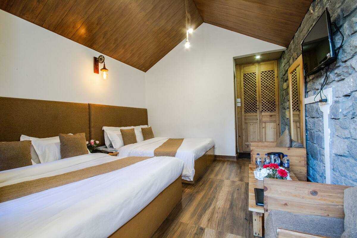 Habitación de hotel con dos camas, techo de madera, pared de piedra y televisor montado en la pared.
