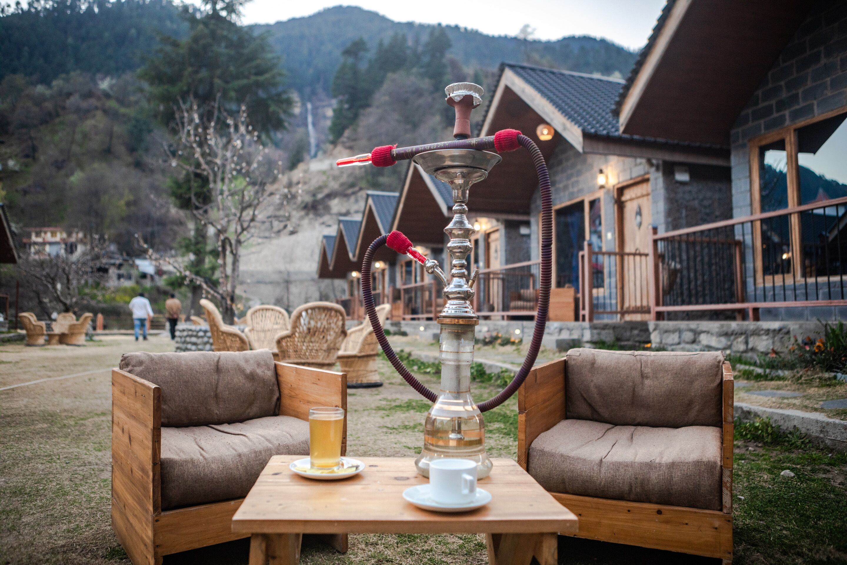 A wooden table with a hookah, a glass of beer, and a white cup. There are two wooden sofas and wicker chairs in the background.