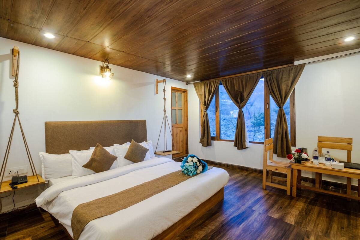 Un dormitorio con techo de madera, una cama con ropa de cama blanca, una mesita de noche de madera, un escritorio con silla y una ventana con cortinas.