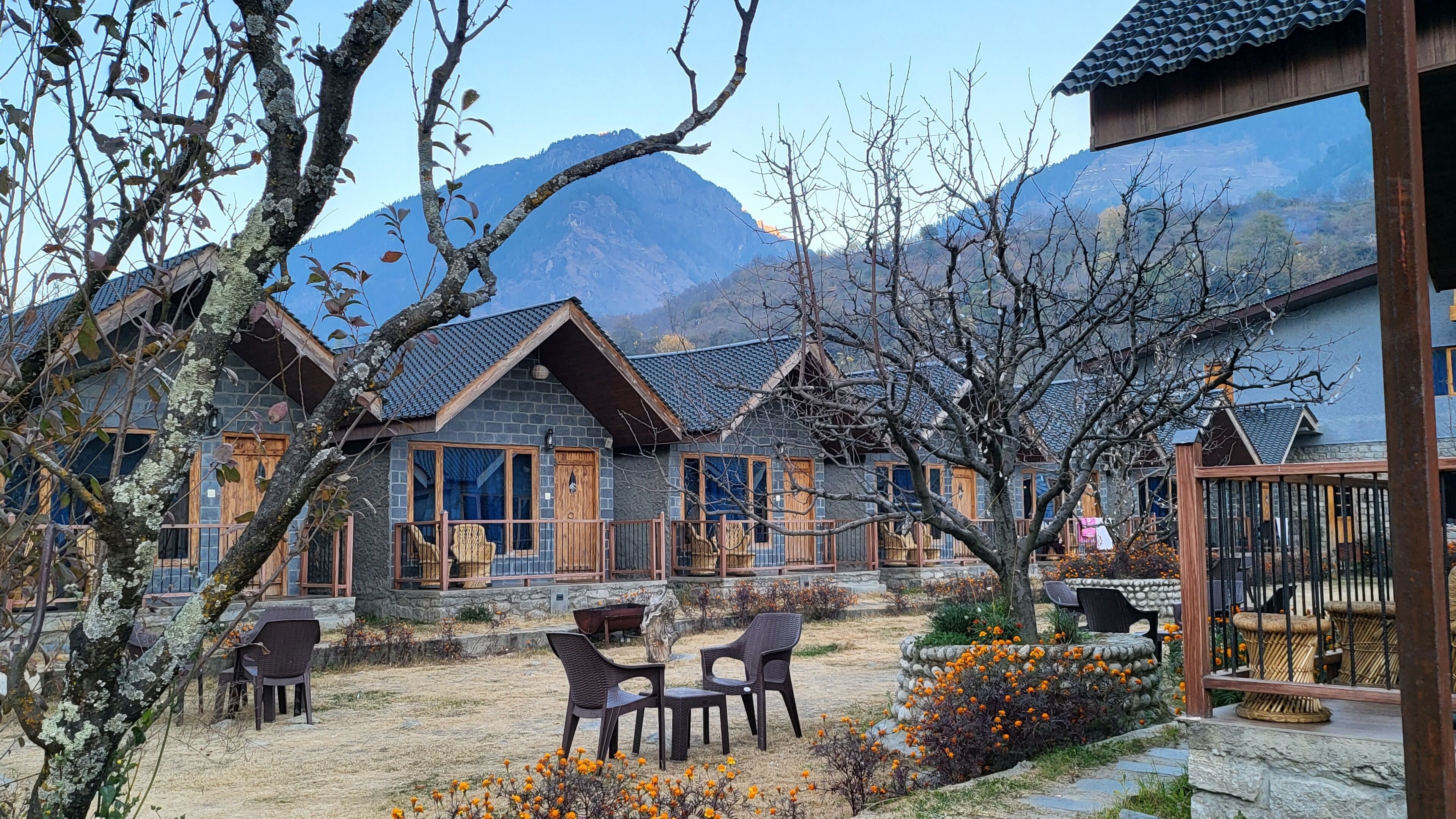 Un bâtiment traditionnel en pierre, doté d’un balcon en bois, entouré d’un jardin où poussent des fleurs d’oranger et offrant une vue sur les montagnes.