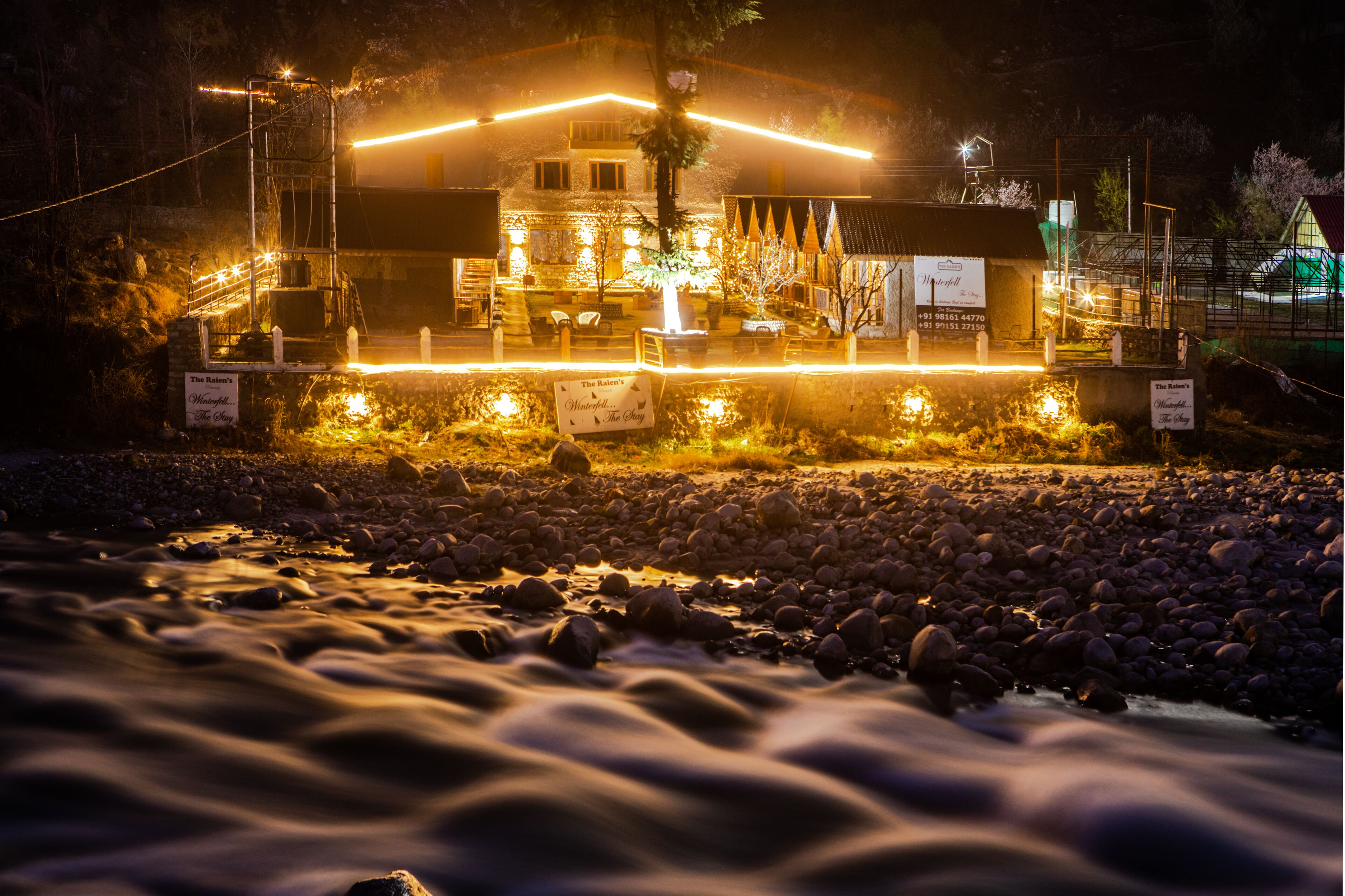 A riverside hotel with a well-lit facade, outdoor seating, and a sign indicating a restaurant.