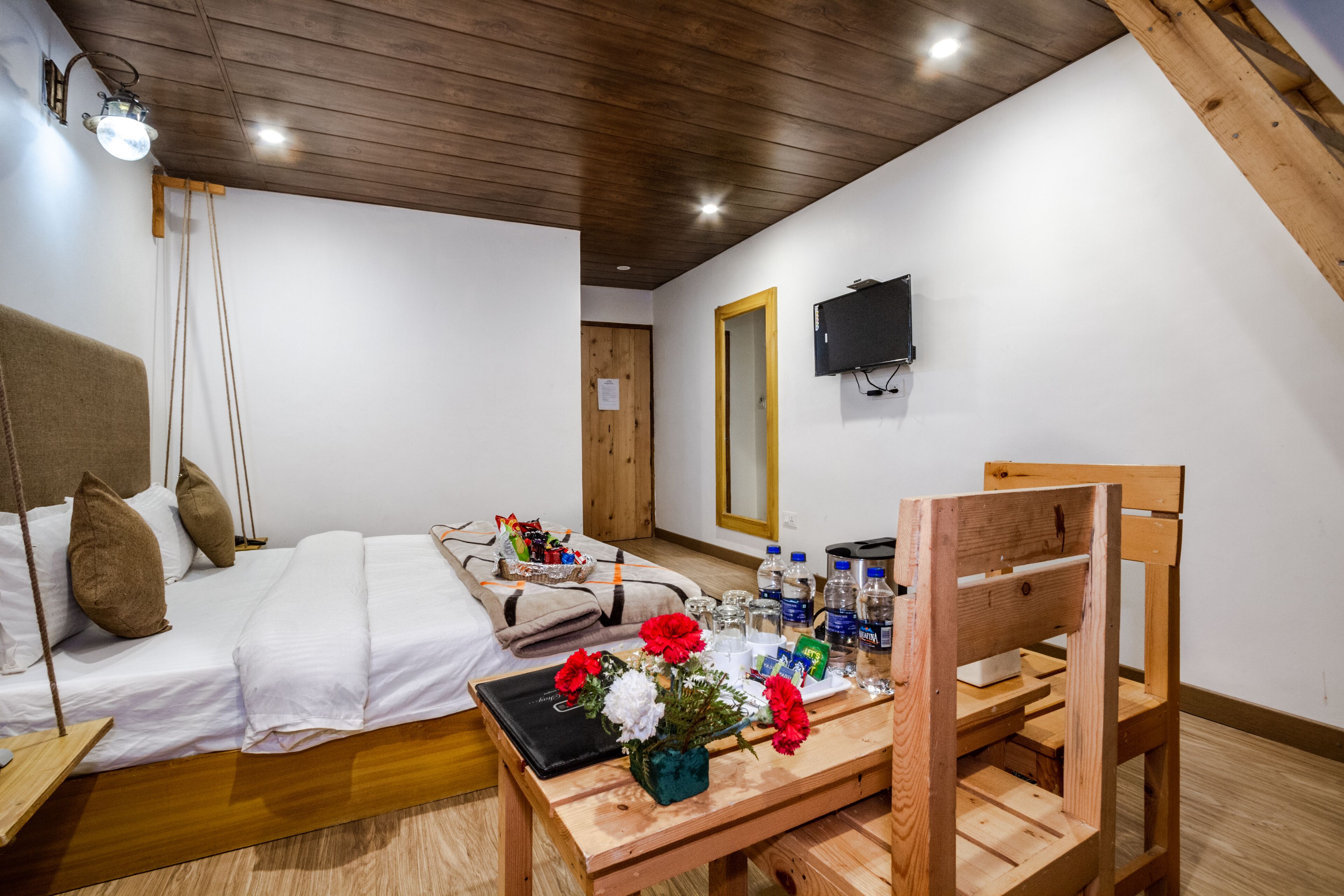 A hotel room with a bed, a wooden chair, a table with a vase of flowers, and a television mounted on the wall.
