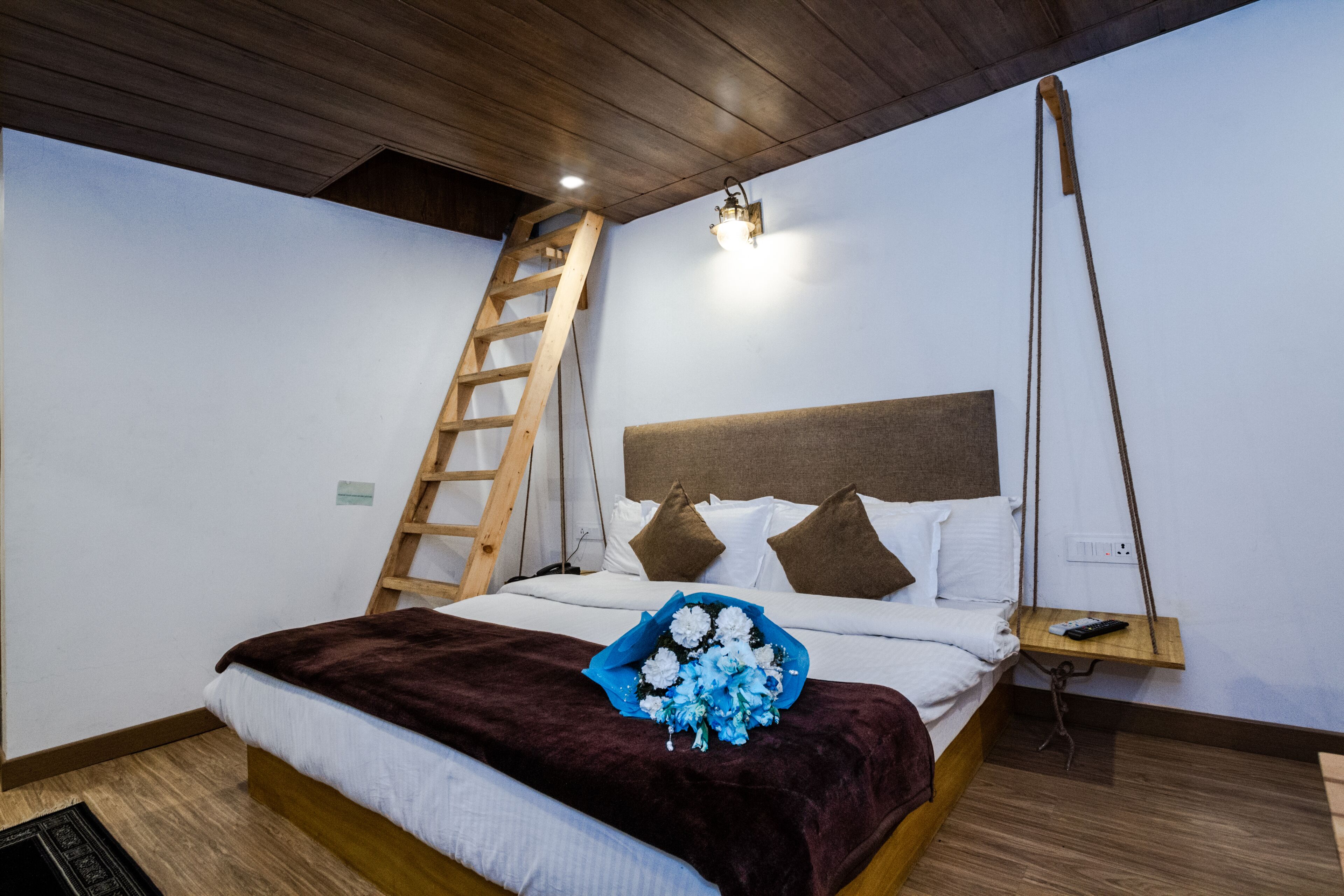 A neatly made bed with a brown blanket, a wooden ladder, a bedside table with a remote control, and a wall-mounted light fixture.