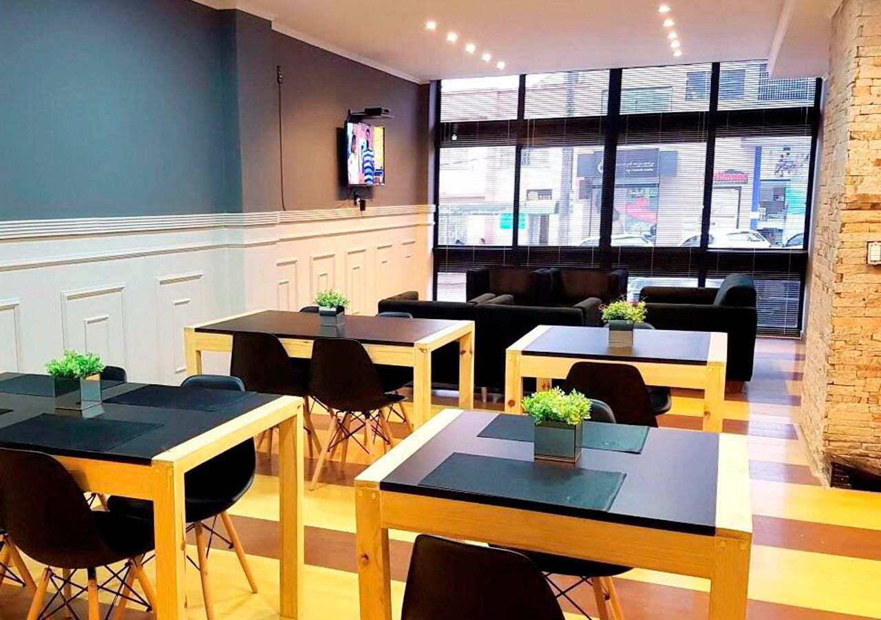 Una cafetería moderna con mesas de madera y sillas negras, amplios ventanales con vistas a la ciudad y un televisor en la pared.