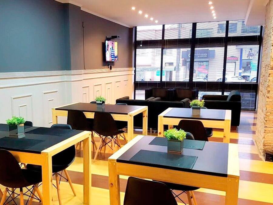 Una cafetería moderna con mesas de madera y sillas negras, amplios ventanales con vistas a la ciudad y un televisor en la pared.