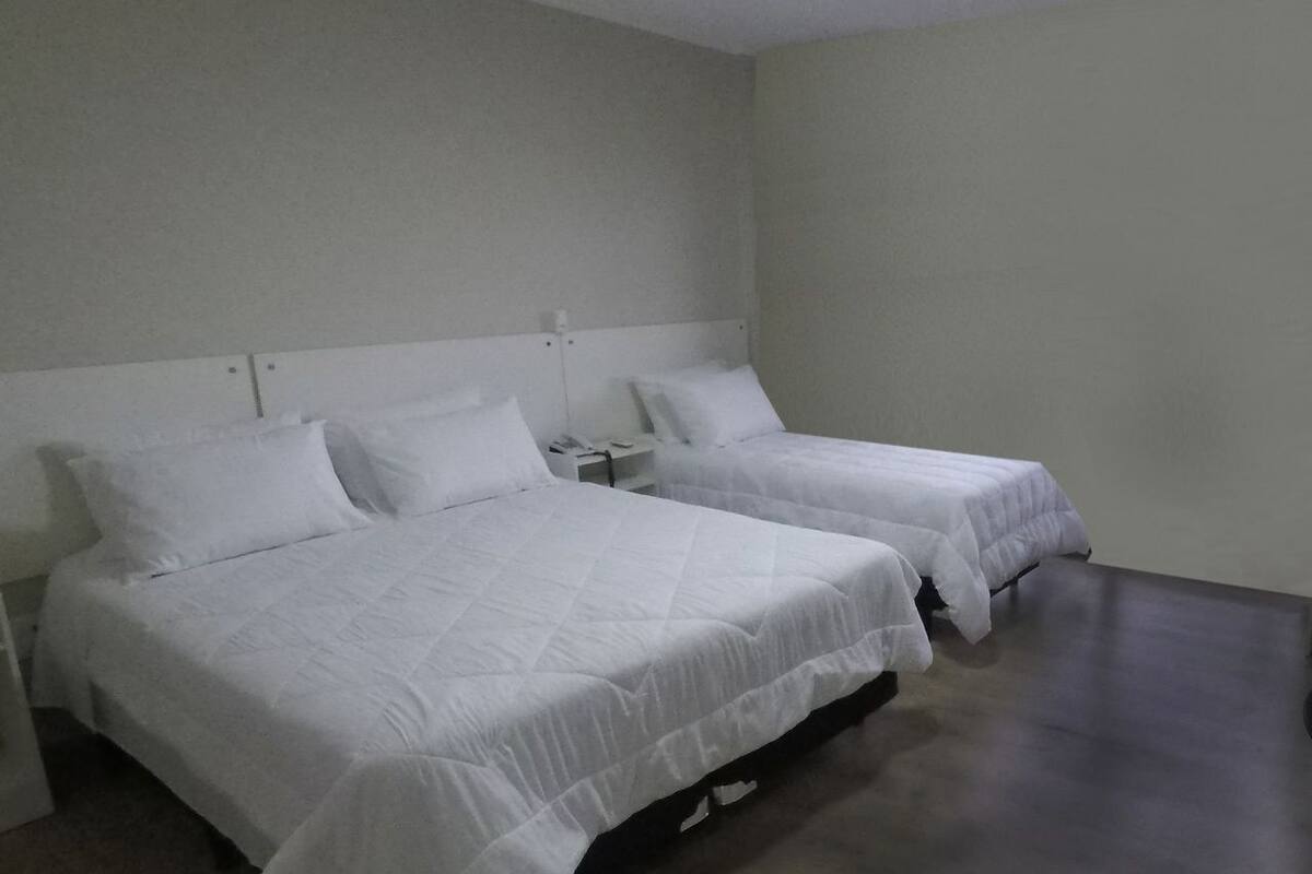 A room with two single beds, each with a white bedspread and matching pillows. There is a nightstand with a lamp between the beds. The room has a simple, clean design with a light-colored wall and a dark floor.