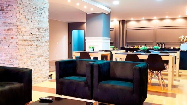 A modern hotel lobby with a brick accent wall, seating area with dark sofas, a central dining table, and a kitchen area in the background.