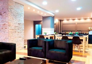 A modern hotel lobby with a brick accent wall, seating area with dark sofas, a central dining table, and a kitchen area in the background. - Hayer Hotel (Erechim)