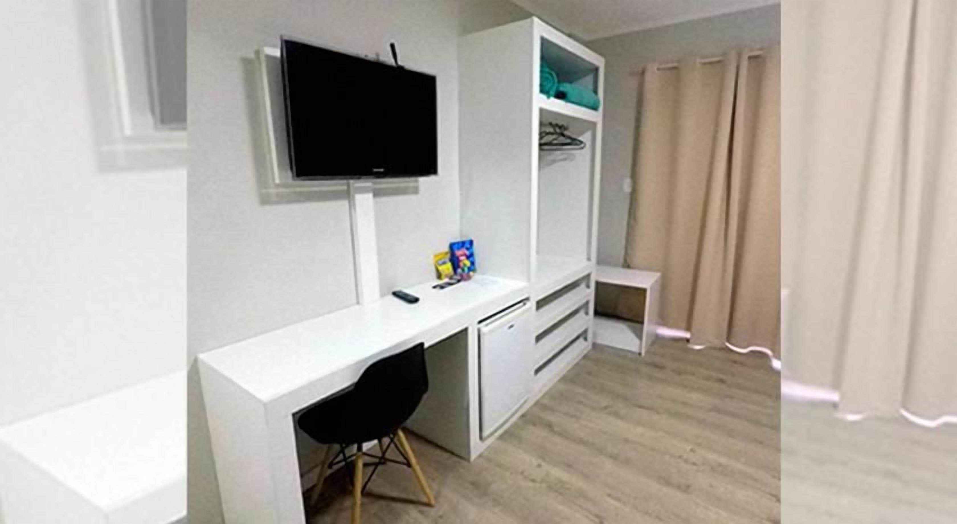 A room with a white desk, a television mounted on the wall, a small seating area, and a window with curtains.