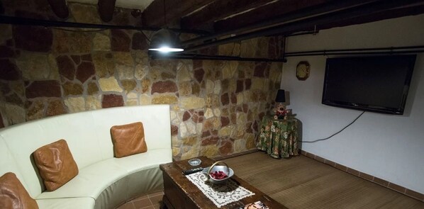 A living room with a stone wall, a white sofa with brown cushions, a wooden coffee table with a bowl of fruit, and a television on the wall.