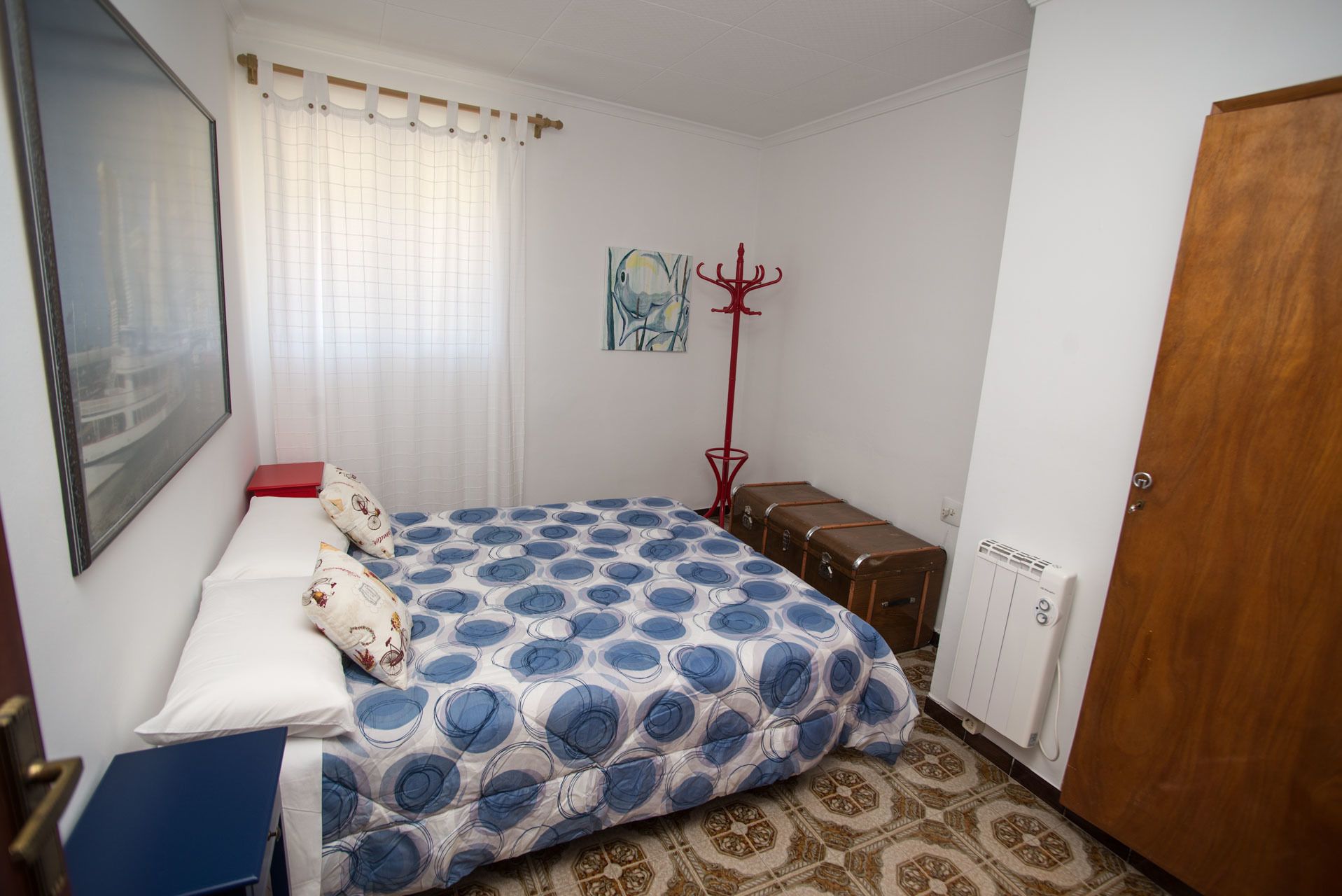 Un dormitorio con una cama, un perchero rojo, una maleta, un radiador y una puerta.