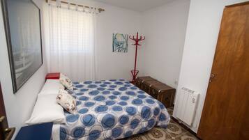 Un dormitorio con una cama, un perchero rojo, una maleta, un radiador y una puerta.