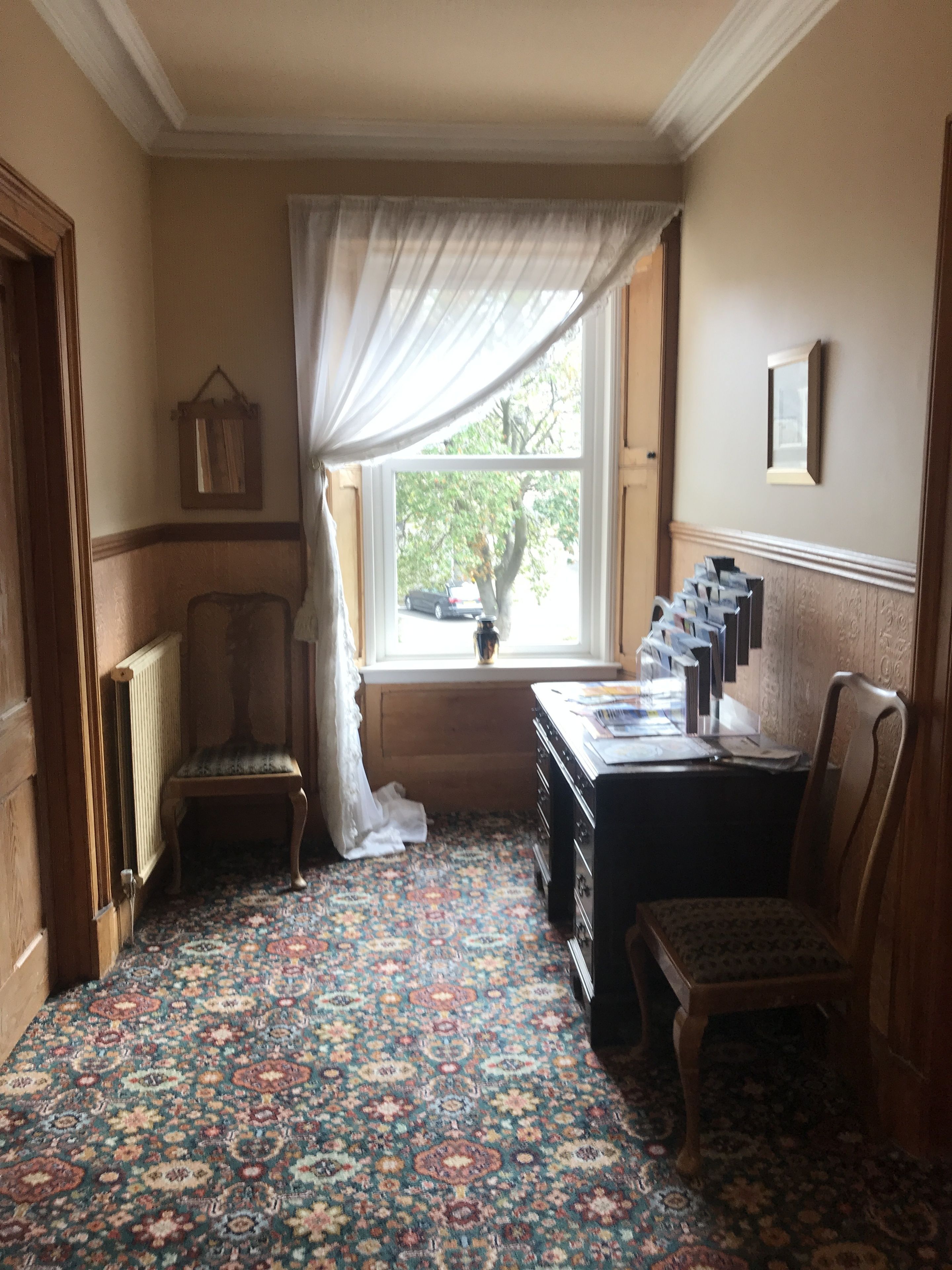 A room with a patterned carpet, a window with a view of trees, a chair, a table with papers, and a radiator.