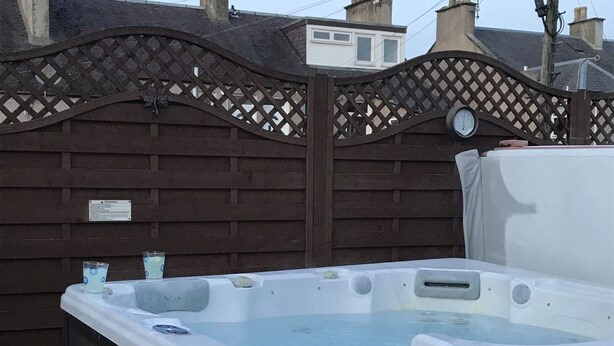 A hot tub in a backyard with a wooden fence and houses in the background. - Dunclutha Guest House (Leven)