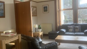 A living room with a black leather sofa, a wooden coffee table, and a dining table with chairs. There is a chandelier hanging from the ceiling, a radiator, and a window with a view of trees. - Dunclutha Guest House (Leven)
