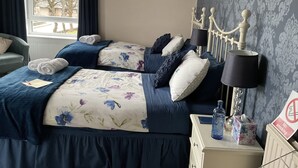 A hotel room with two beds, dark blue bedding, floral patterns, a bedside table with a lamp, a water bottle, and a tissue box. - Dunclutha Guest House (Leven)