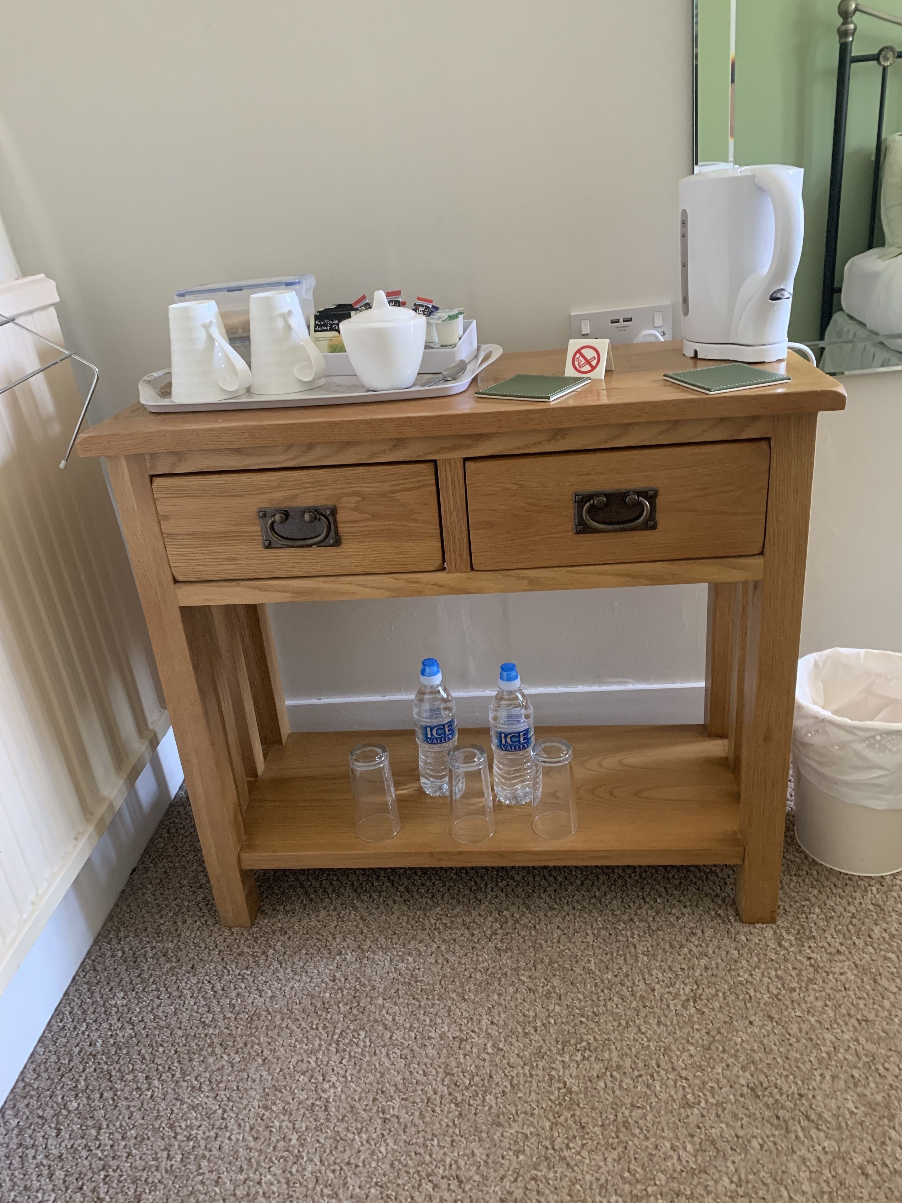 A wooden desk with two drawers, a tray with white cups and a teapot, a notebook, a water bottle, and a coffee maker.