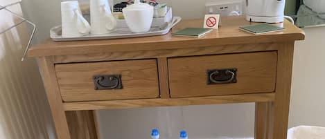 A wooden desk with two drawers, a tray with white cups and a teapot, a notebook, a water bottle, and a coffee maker.