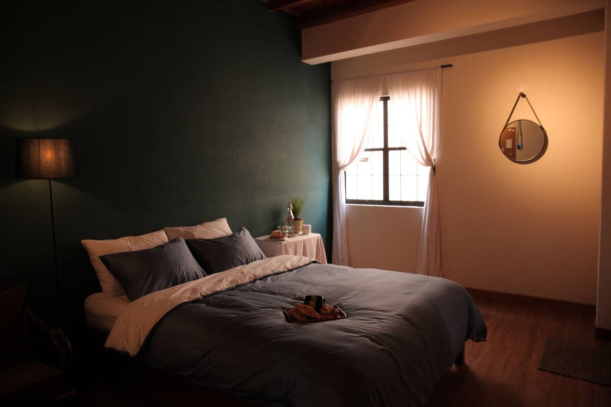 A bedroom with a bed, a bedside table with a lamp, a window with curtains, and a wall clock.