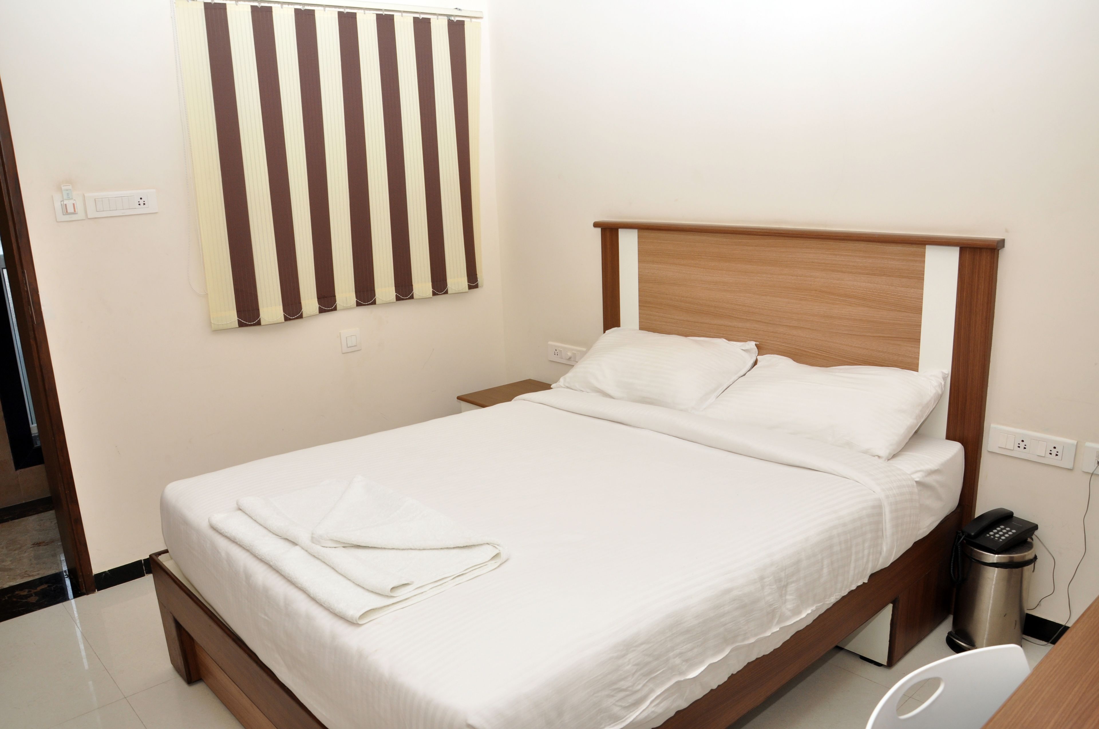 Een net opgemaakt bed met witte lakens, een houten hoofdbord en een nachtkastje met een prullenbak.