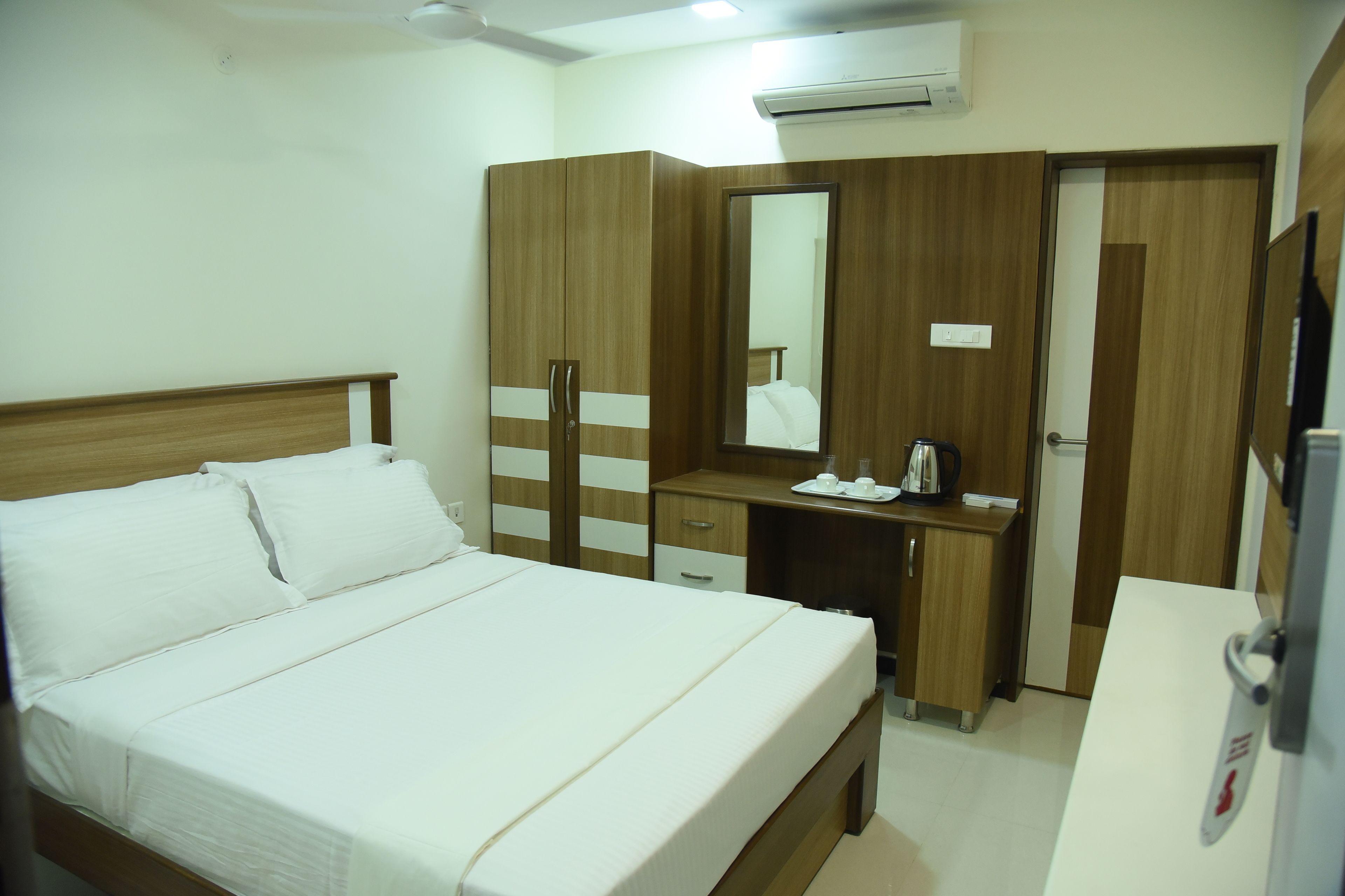 A hotel room with a bed, a wooden wardrobe, a desk with a mirror, and a white bedside table.