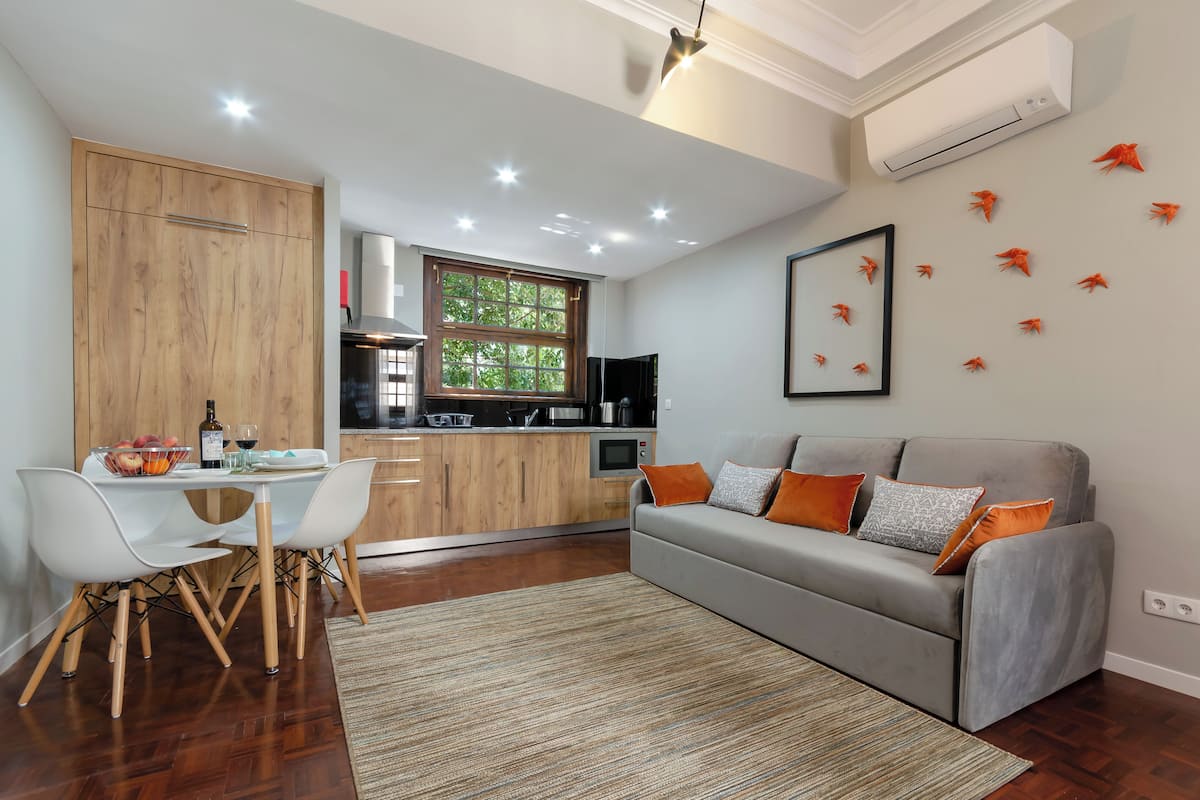 A modern kitchen with wooden cabinets, a dining area with white chairs, and a grey sofa with orange cushions.