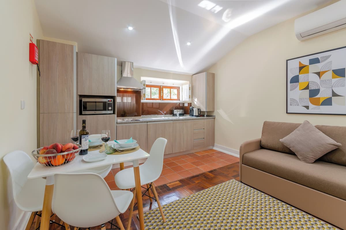 A modern kitchen with a microwave, stove, and sink. There is a dining table with chairs and a fruit bowl. A sofa and a patterned rug are in the living area.