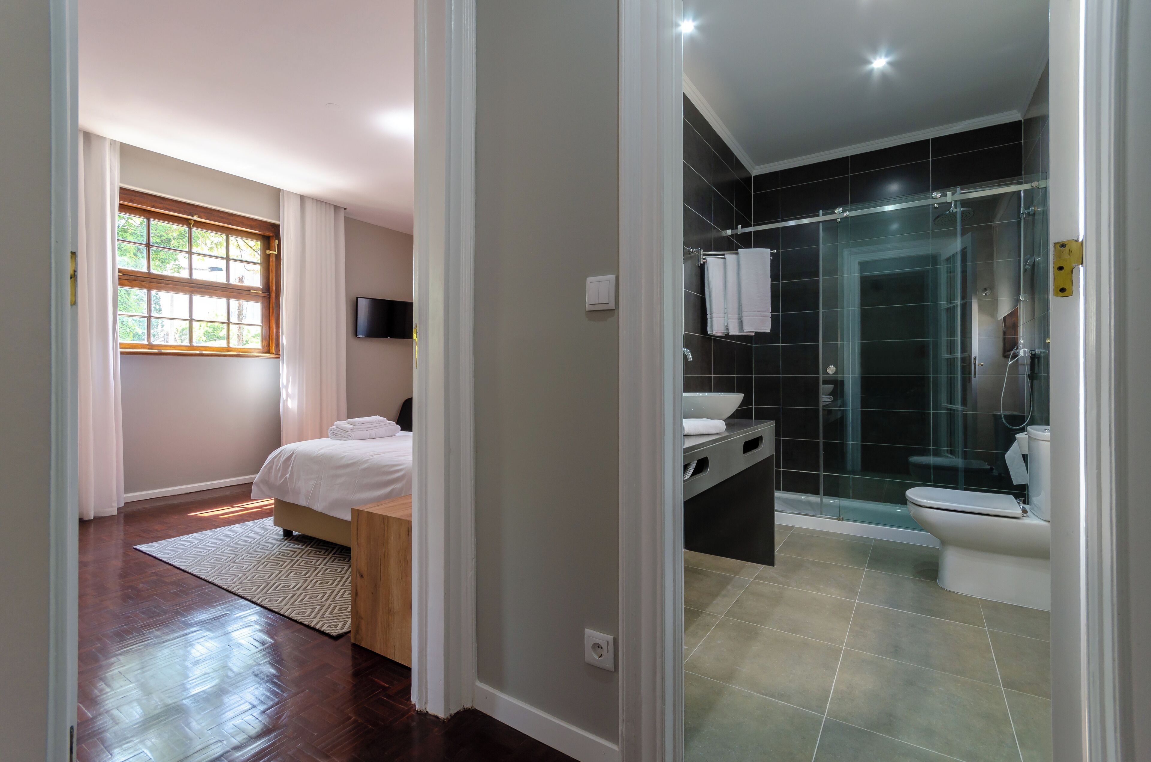 Ein modernes Hotelzimmer mit Holzboden, einem Bett, einem Fenster mit Vorhängen, einem Badezimmer mit Duschkabine und einer Toilette.