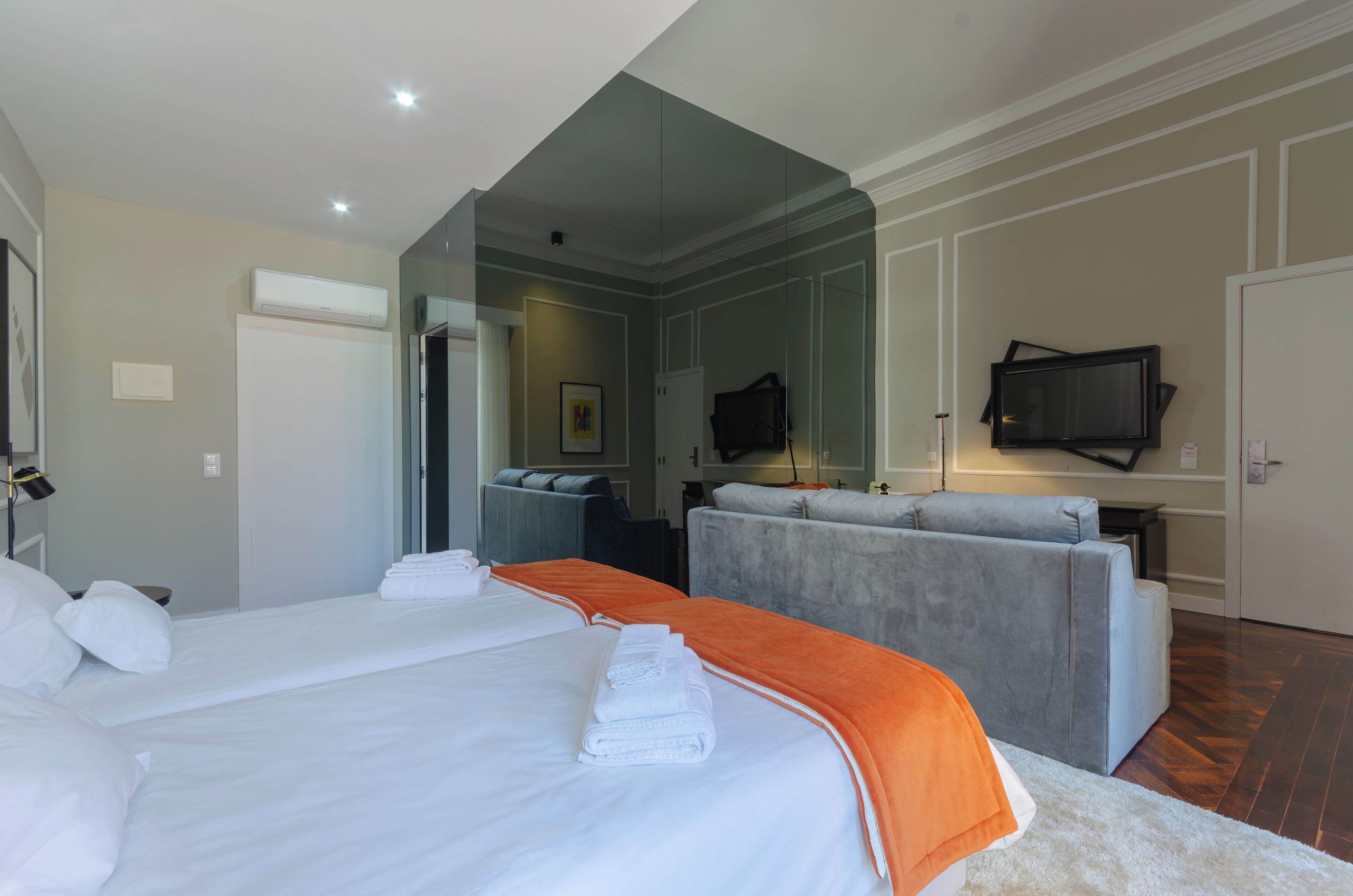 Una habitación de hotel con dos camas, un sofá gris, un televisor y un espejo.