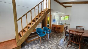 Een houten trap leidt naar een slaapkamer, een keuken met een spoelbak en fornuis, en een eethoek met stoelen en een tafel.