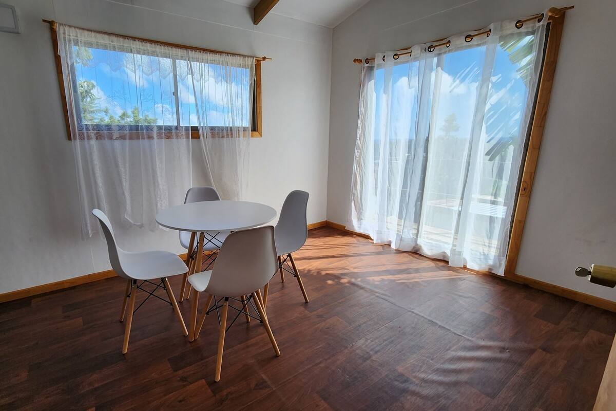 A room with a round table and four chairs, wooden flooring, and large windows with sheer curtains.