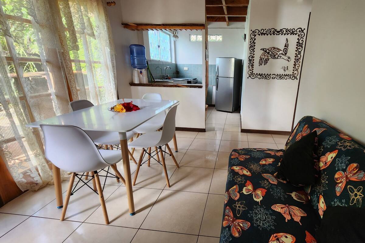A dining area with a white table and chairs, a kitchen with a refrigerator, and a patterned sofa.