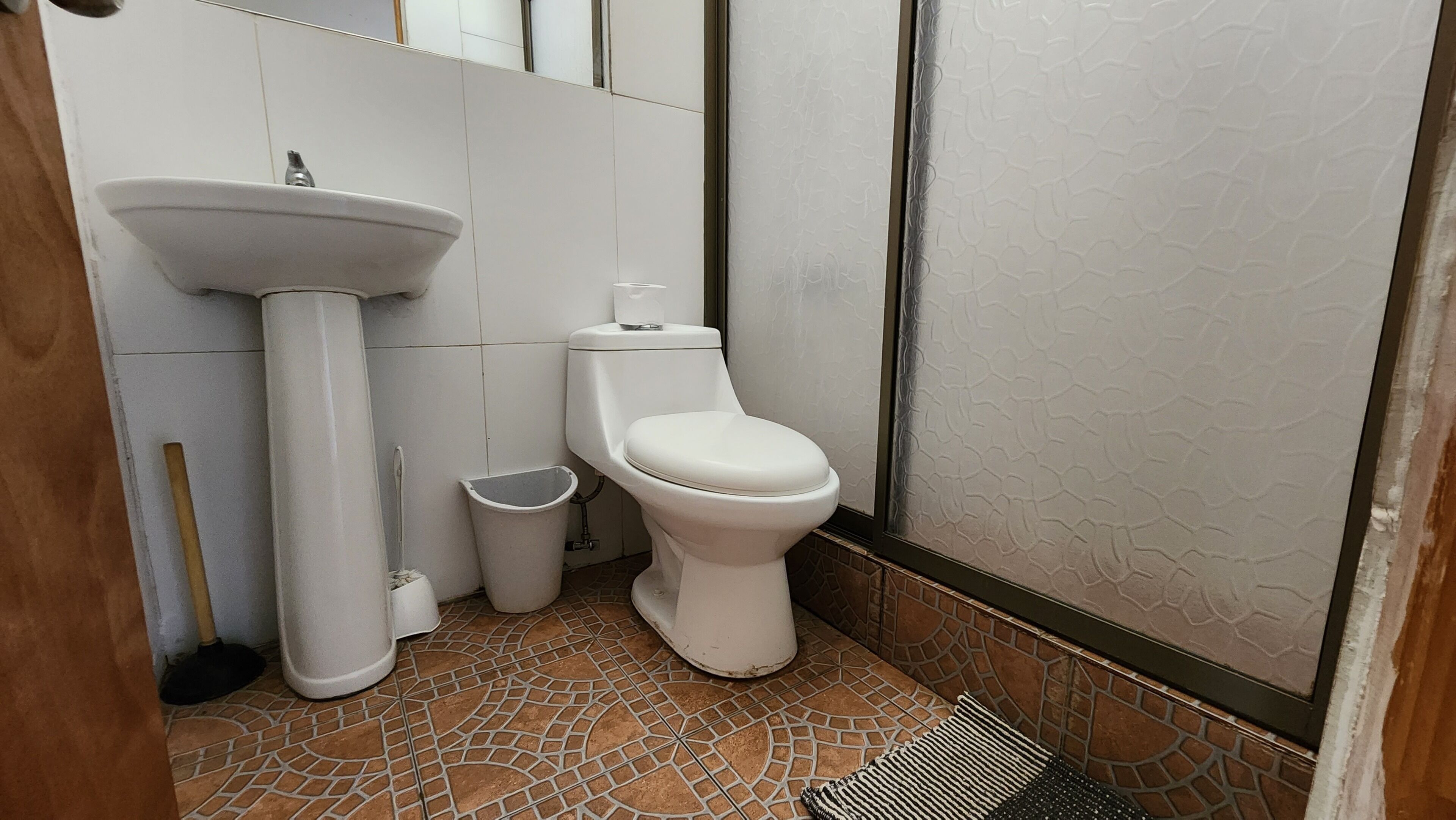 Une salle de bain avec un lavabo sur pied blanc, des toilettes et une poubelle. Le sol est recouvert de carreaux formant un motif.