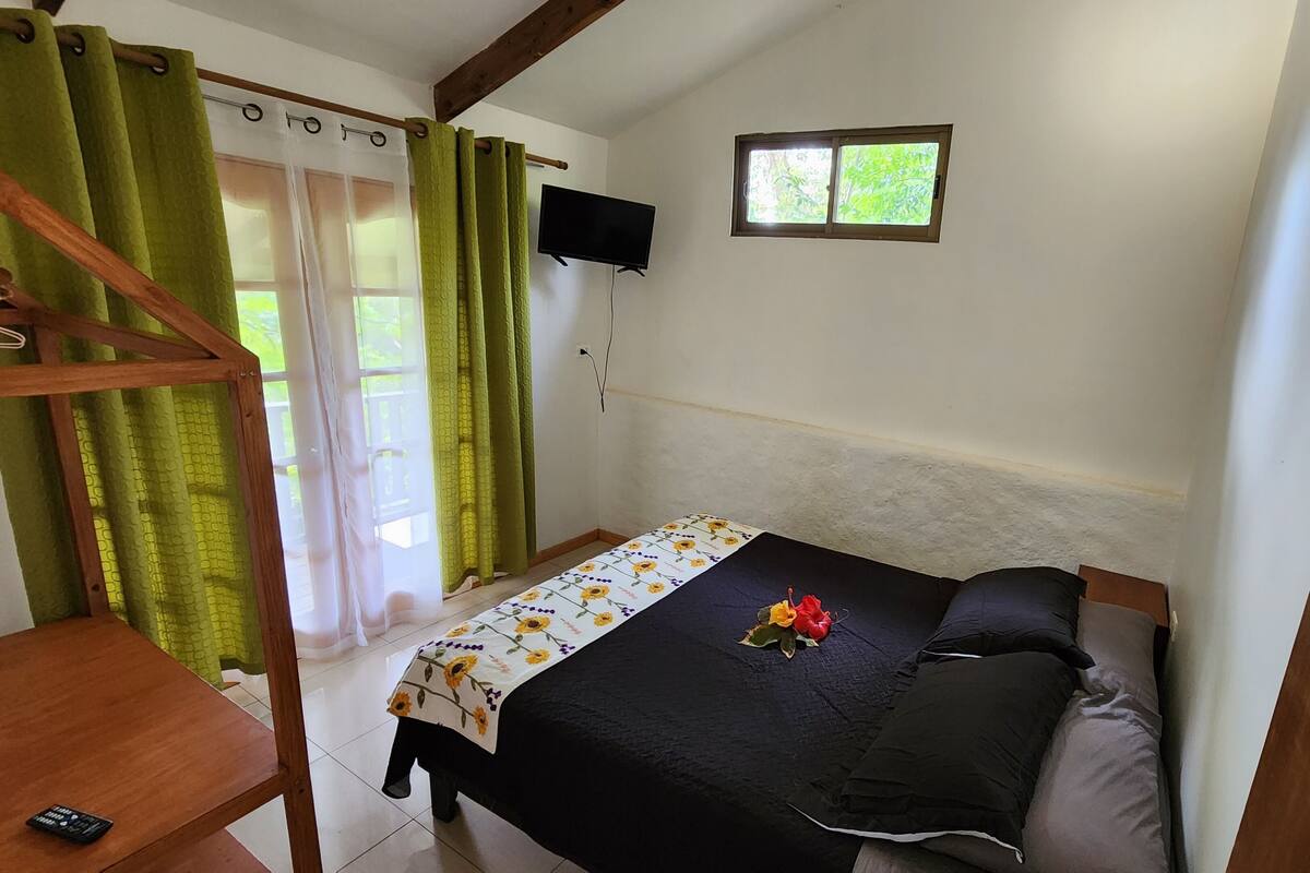 A small bedroom with a bed, a TV mounted on the wall, a window, and a wooden ceiling beam.