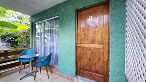 Ein Zimmer mit Holztür, einem Fenster mit Vorhang, einem kleinen Tisch mit Stühlen und einem Balkon mit weißem Gitterzaun.