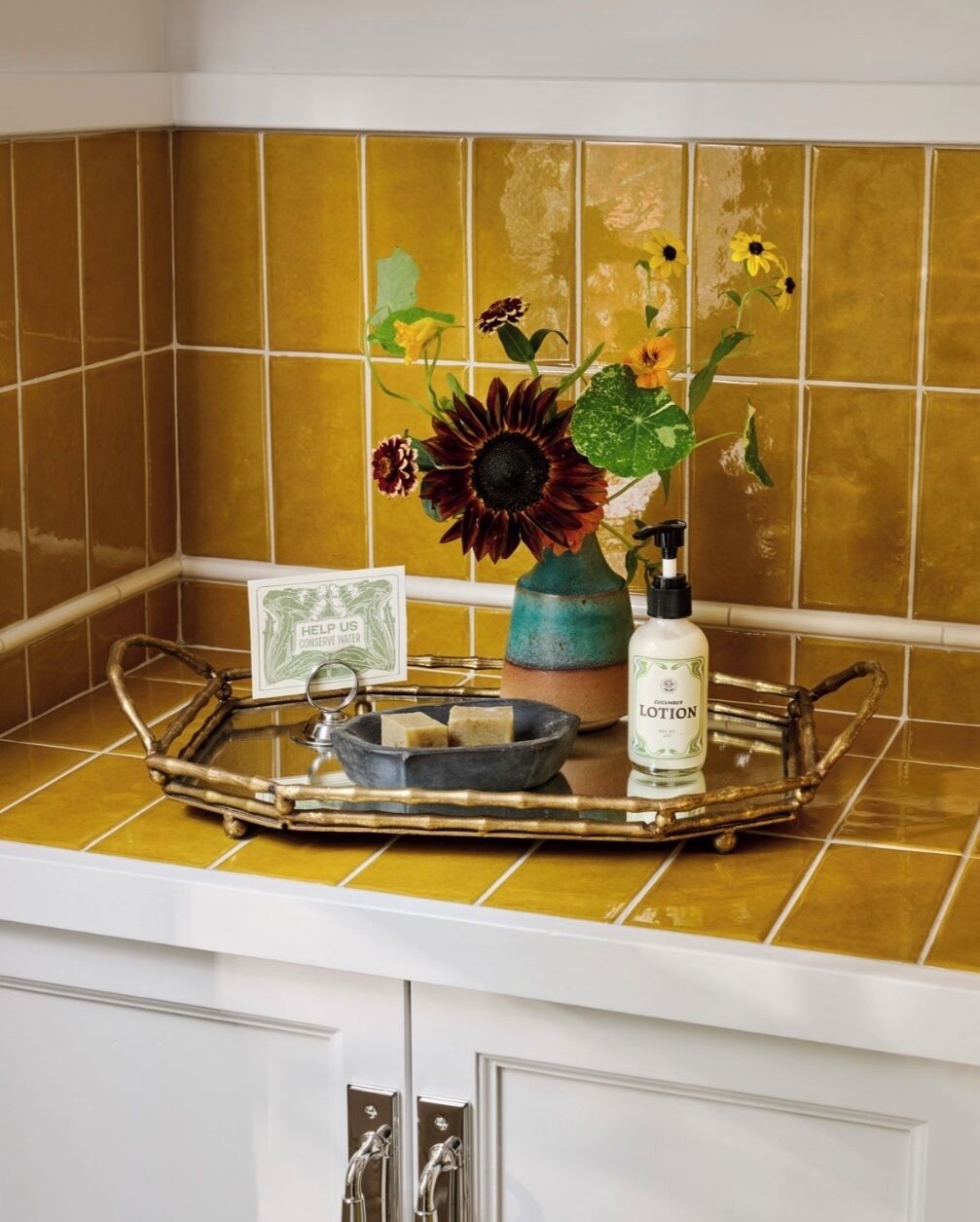 A kitchen counter with a tray holding a vase of flowers, a soap dispenser, and a sign encouraging water conservation.