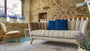 A beige sofa with a blue pillow, a wooden chair, and a stone wall. - Rúa Travesa (Santiago de Compostela)