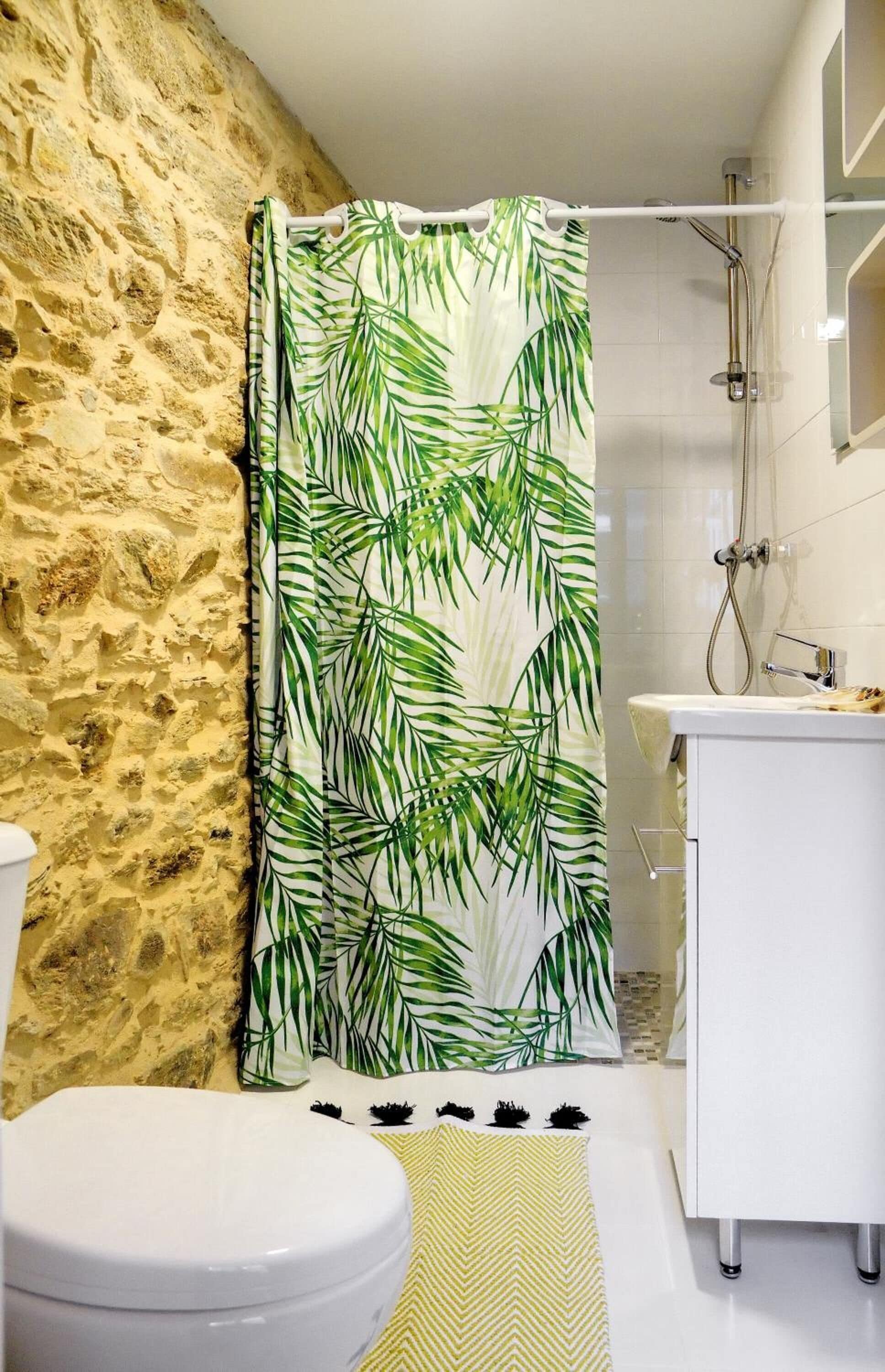 Een badkamer met een stenen muur, een douchegordijn met een bladmotief, een witte wastafel en een toilet.