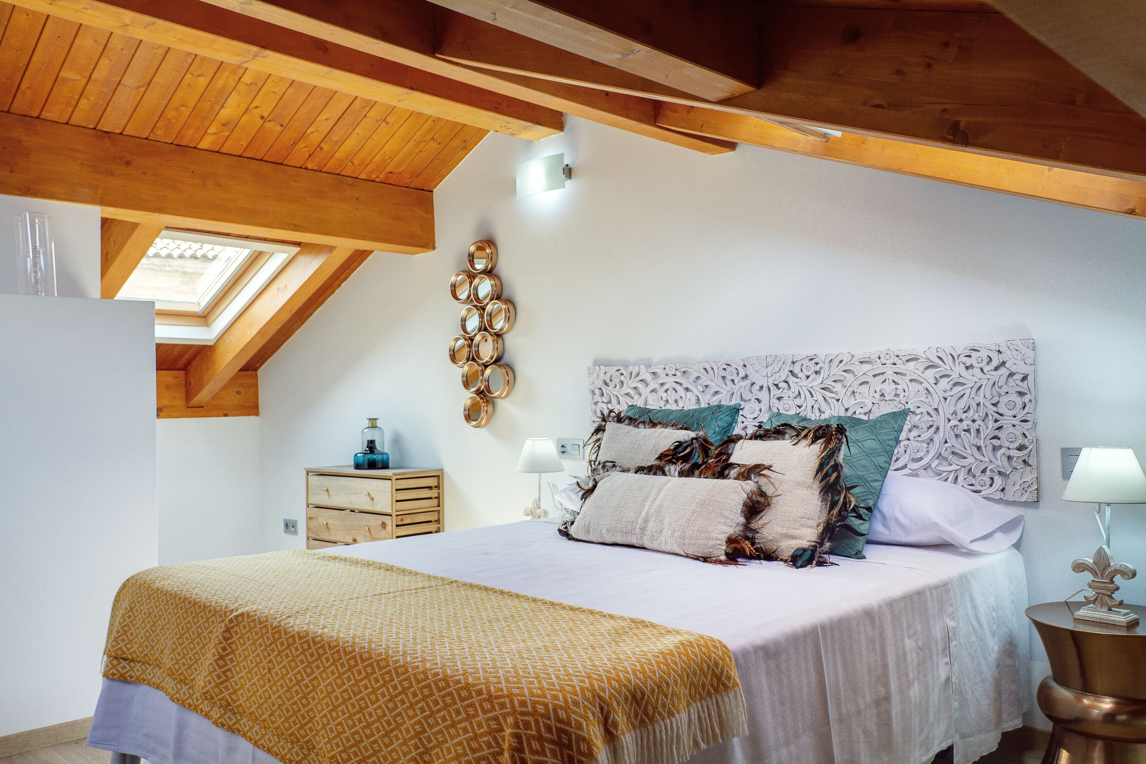 A bedroom with a wooden ceiling, a skylight, a bed with patterned bedding, a bedside table with a lamp, and a wall-mounted mirror.