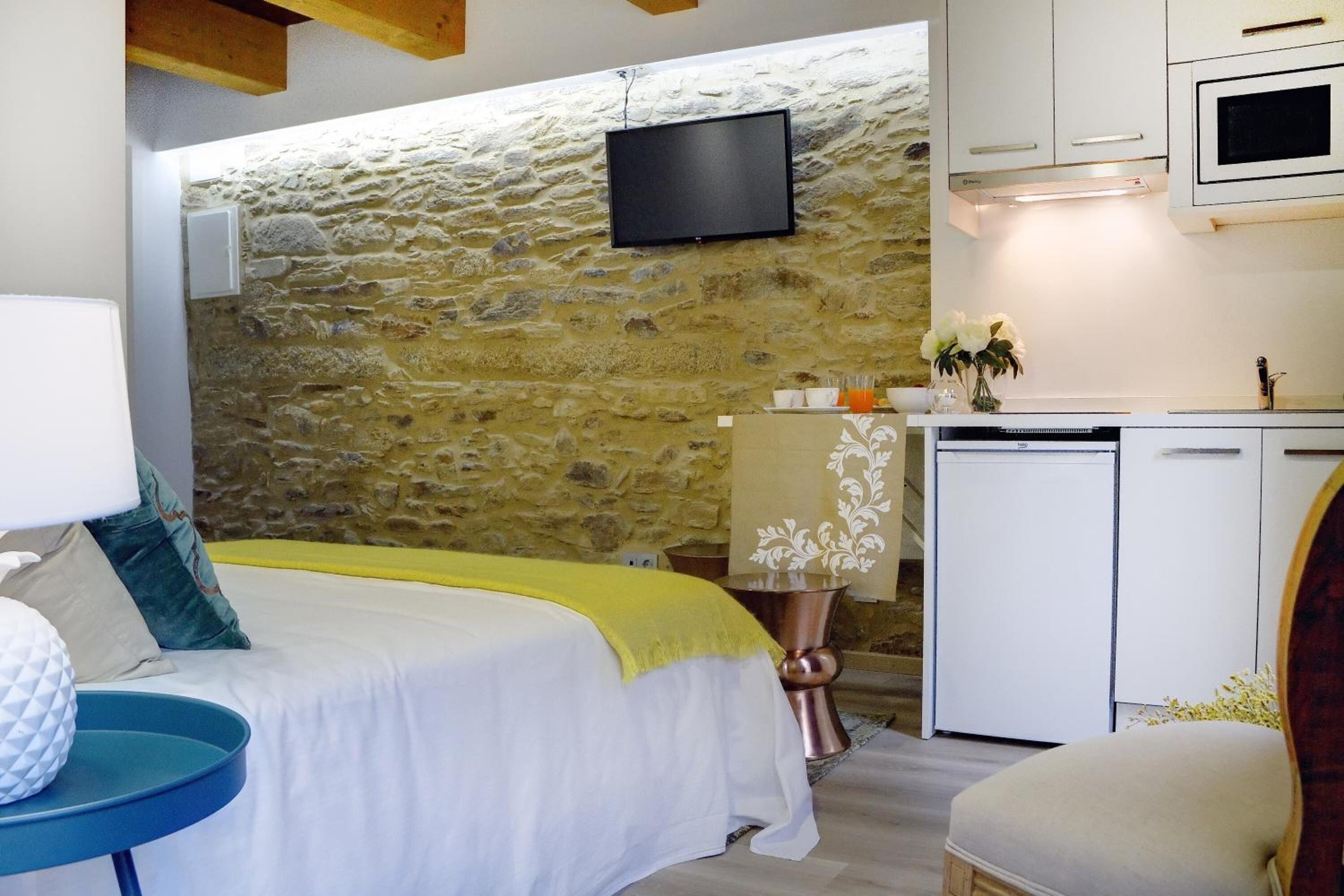 A bedroom with a stone wall, a bed with a yellow blanket, a bedside table with a lamp, and a small kitchenette with a microwave.