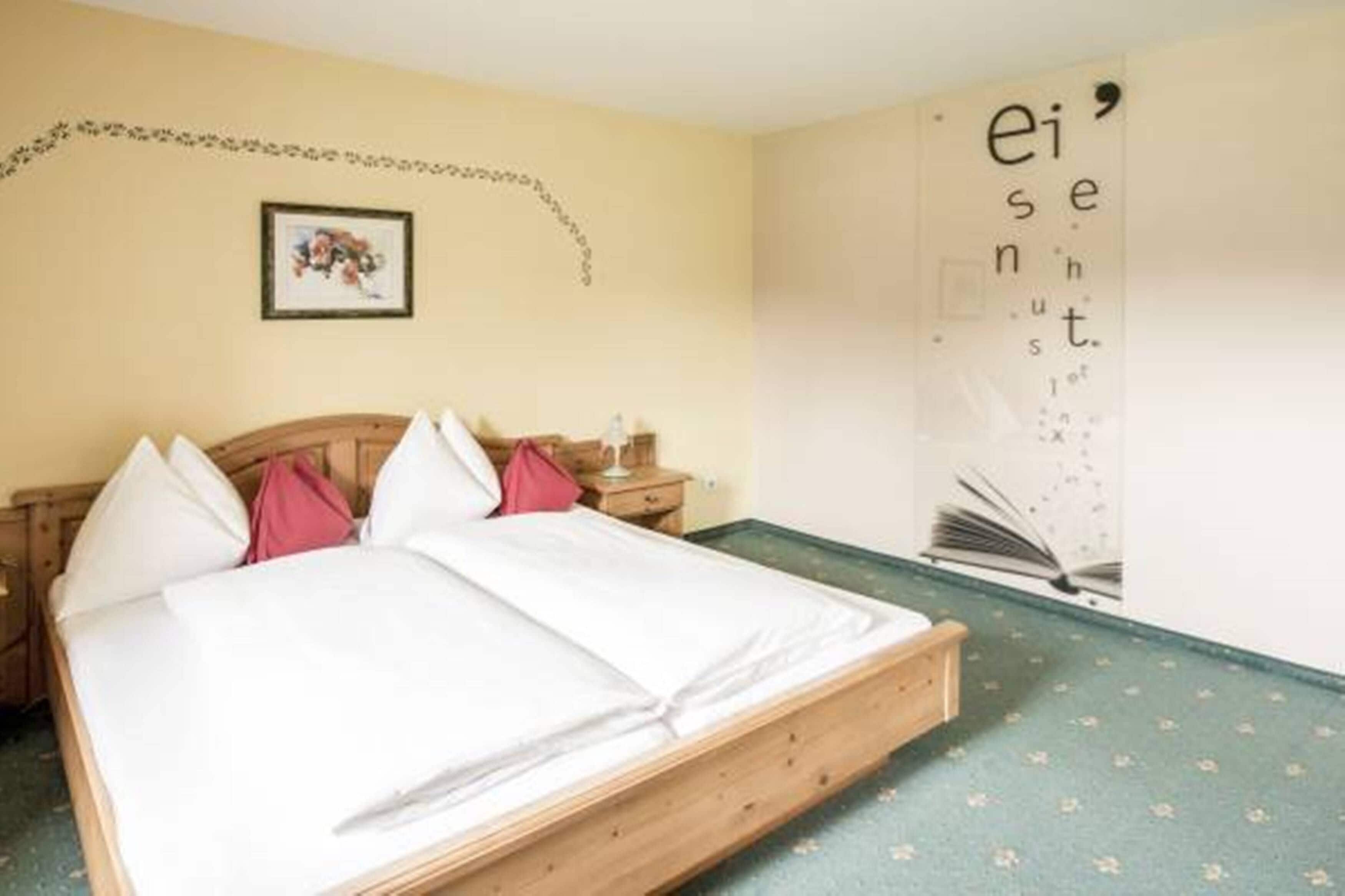 A bedroom with a wooden bed, white bedding, a small bedside table, and a wall with a partial text and a framed picture.