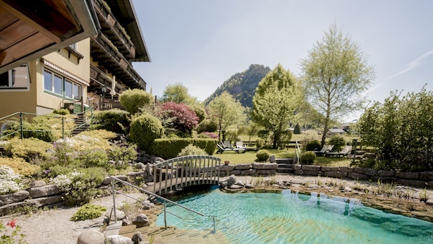 Un área de piscina con un puente de madera, rodeada de abundante vegetación y una montaña al fondo.
