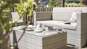 Un espacio de descanso al aire libre con muebles de mimbre, un sofá acolchado y una mesa con superficie de vidrio, frente a un fondo de vegetación y una valla de madera.