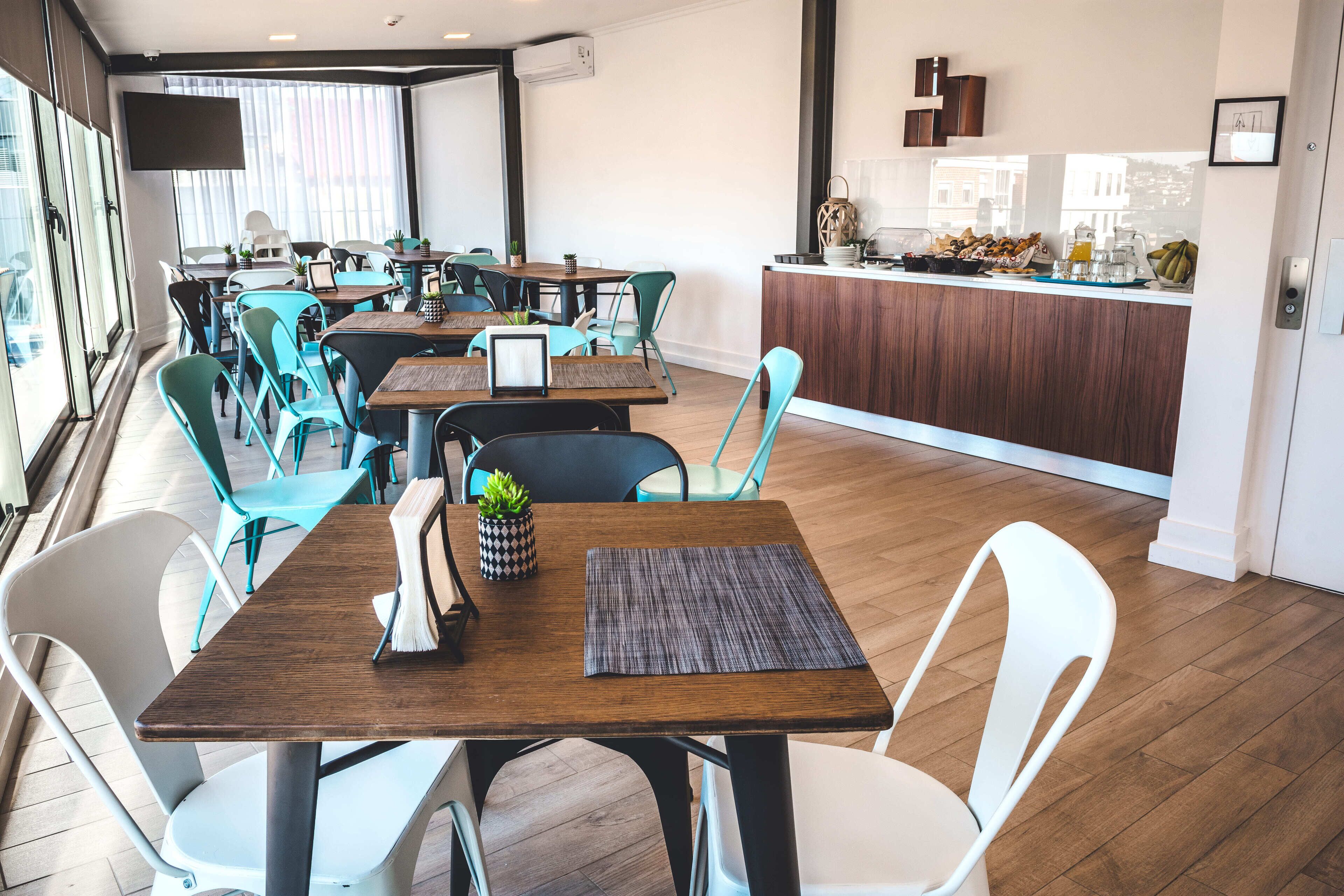 A modern dining area with wooden tables and chairs, a kitchen counter with breakfast items, and large windows providing natural light.