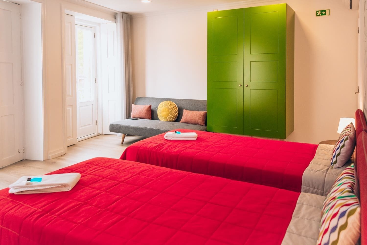Habitación de hotel con dos camas, un armario verde, un sofá gris y una ventana con cortinas.