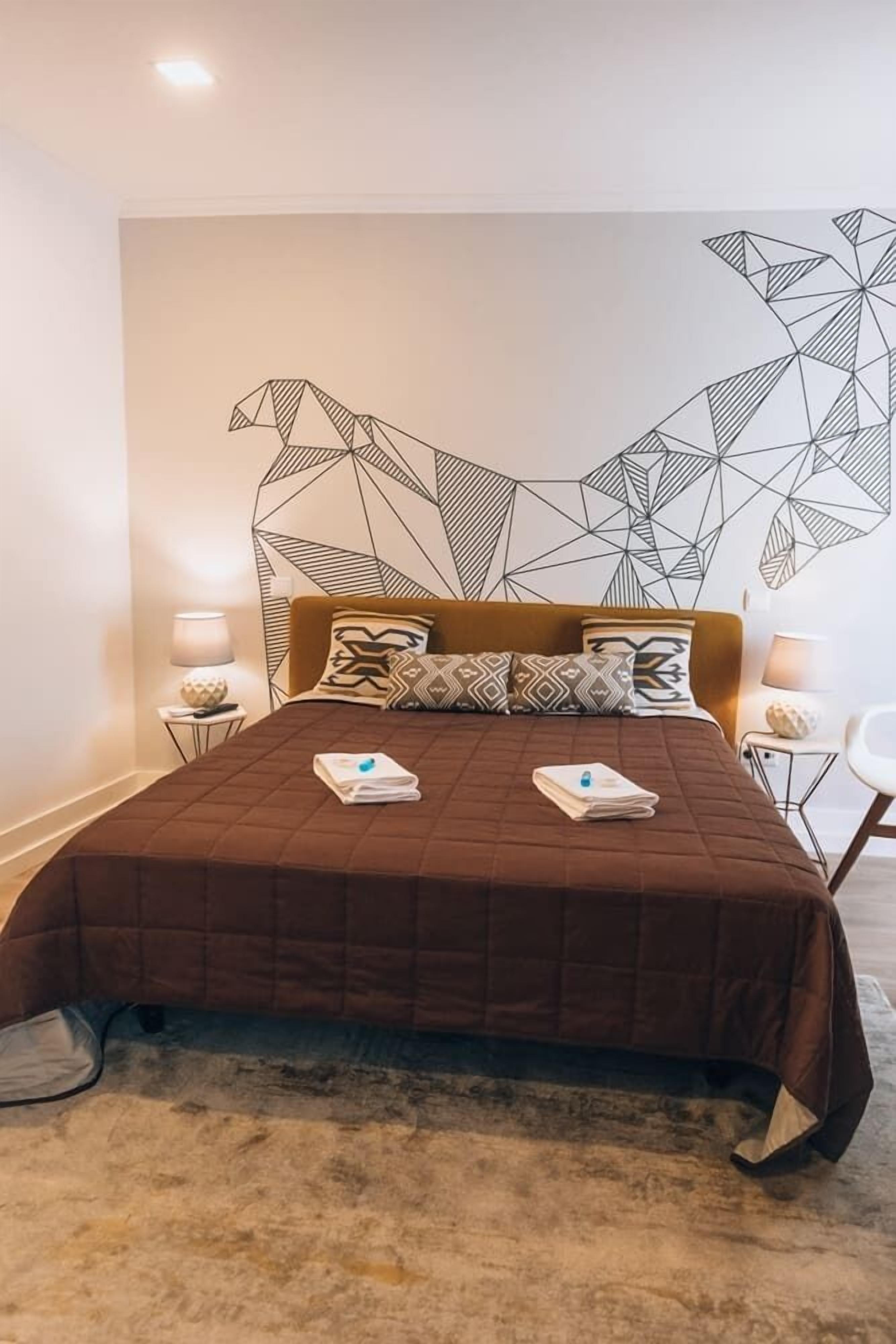 A modern hotel room with a geometric wall art, a brown quilted bedspread, and two bedside lamps.