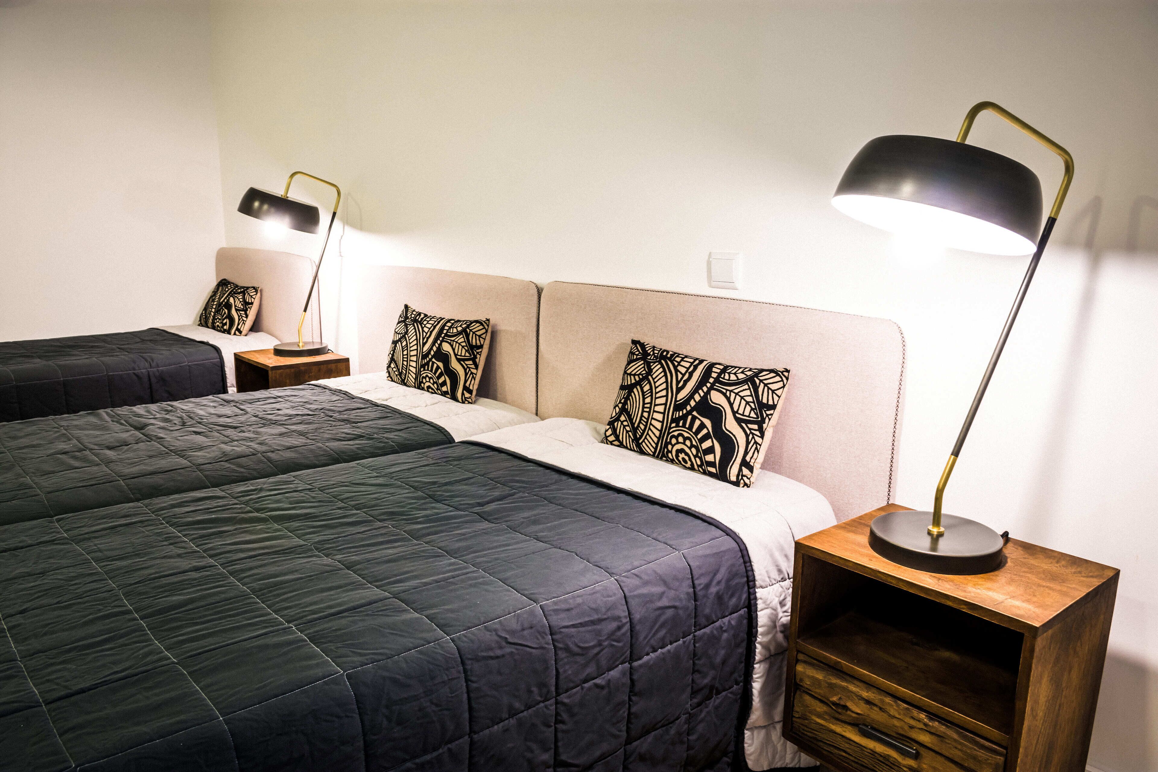 Habitación de hotel con dos camas, cada una con un estilo diferente de ropa de cama, una mesita de noche con lámpara y un pequeño cajón de madera.
